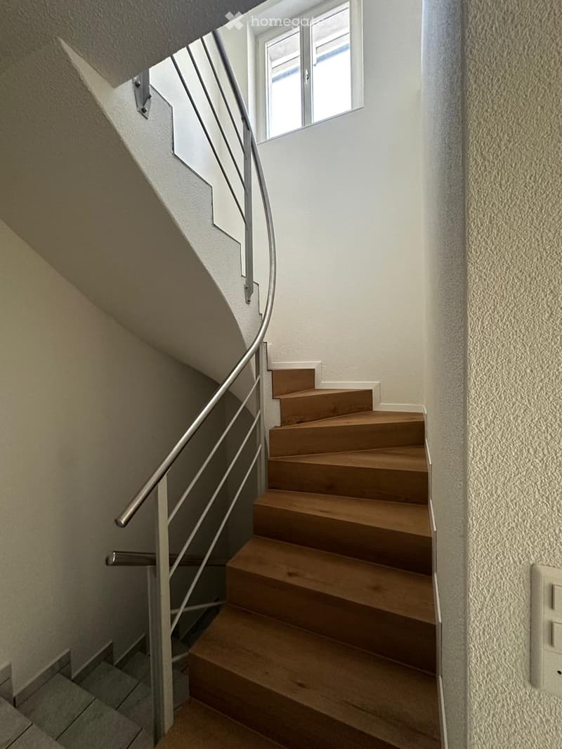 Wooden staircase with metal railing, white walls, and a window at the top landing.