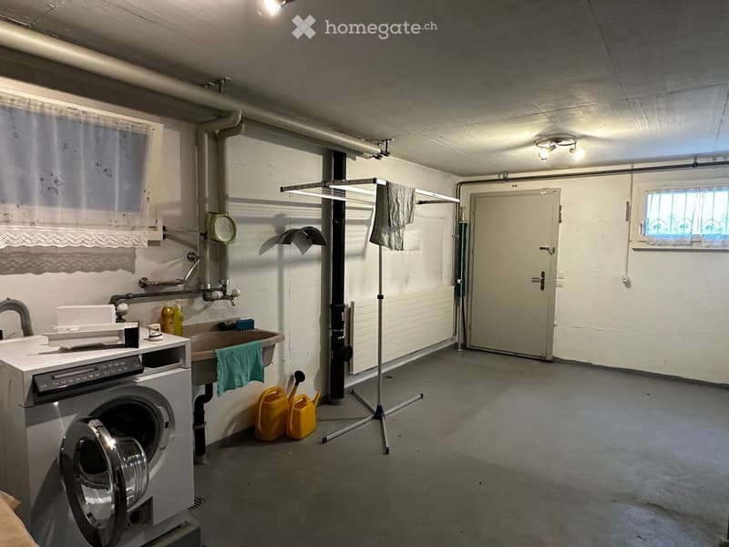 The image shows a small, functional bathroom with a washing machine, sink, and shower area. The space includes a towel rack with a towel, a shower curtain, and a shower stall with a hose and handheld showerhead. There are also cleaning supplies and a smal