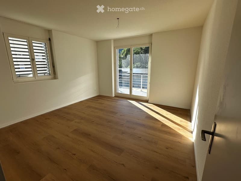 Empty room with wooden flooring, white walls, large window with blinds, and sliding glass door leading to a balcony.