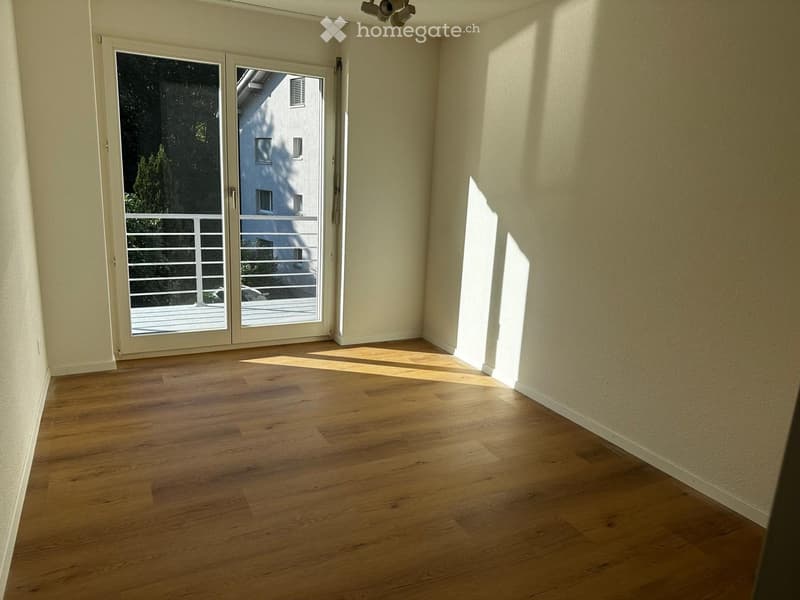 Empty room with wooden flooring, large sliding glass door leading to a balcony, and natural light streaming in.