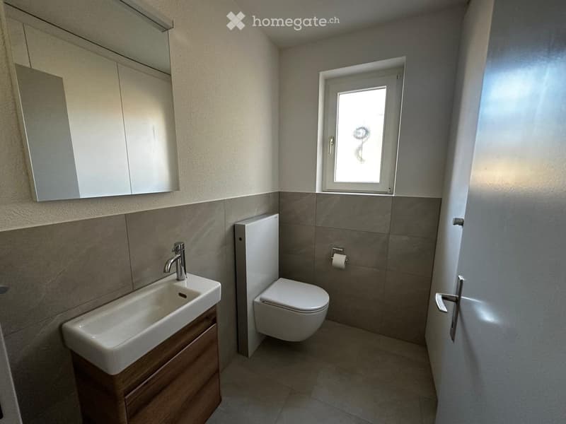 Modern bathroom with a white sink, chrome faucet, toilet, wooden cabinet, and a mirror above the sink. The walls are tiled in a neutral color scheme, and there is a small window providing natural light.