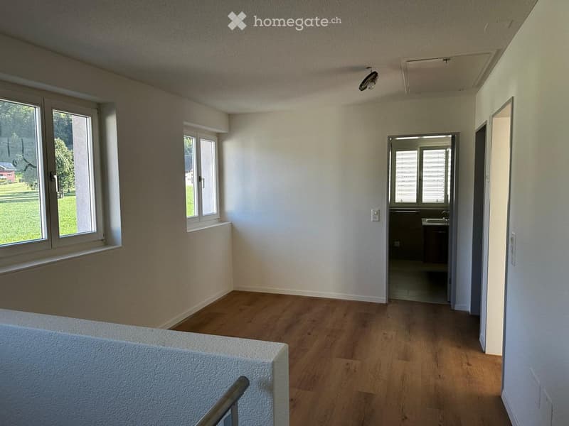 Empty room with wooden flooring, white walls, and large windows. A small kitchen area is visible through an open doorway.