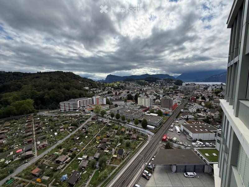 Aerial view of a town with residential buildings, a railway line, and surrounding greenery, with mountains in the background.