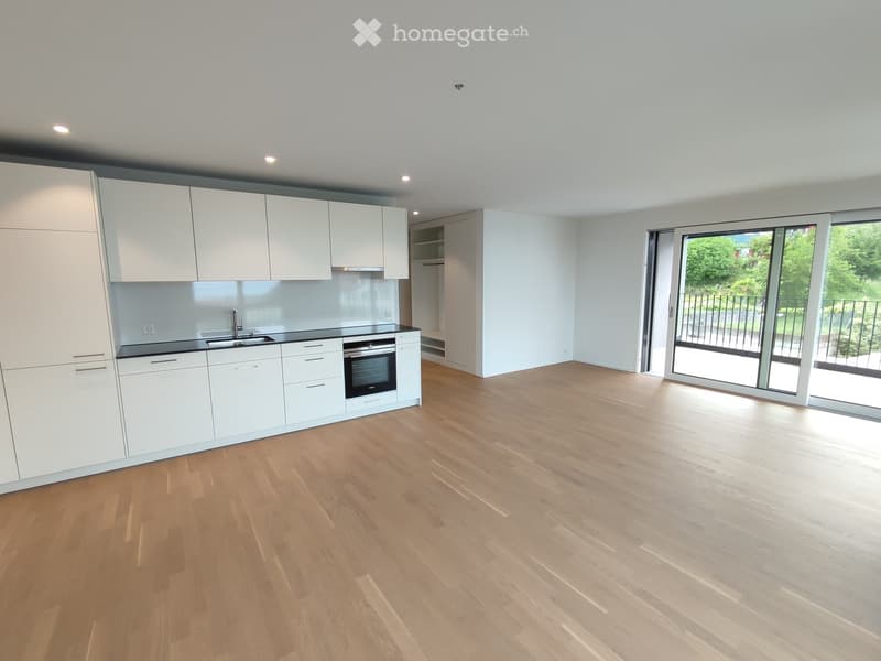 Empty kitchen with white cabinets, black countertops, and a wooden floor. A large sliding glass door leads to a balcony with a view of greenery.