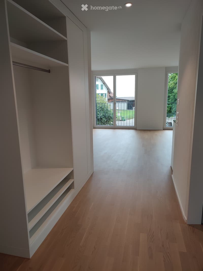 Empty hallway with wooden flooring, built-in shelving on one side, and large windows leading to an outdoor area.