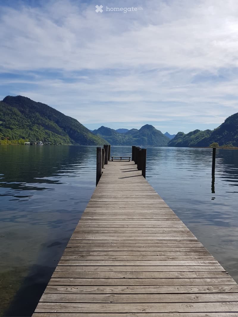 A wooden dock extends into a calm lake surrounded by green, forested mountains under a partly cloudy sky.