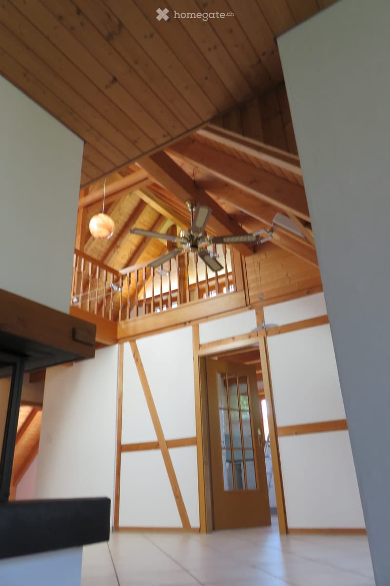 Open, airy hallway with wooden beams and a high, slanted wooden ceiling. Features a wooden railing leading to an upper level and a large open doorway.