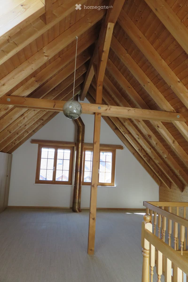 An empty attic space with wooden beams, exposed wooden ceiling, and a staircase leading up, featuring large windows and a hanging light fixture.