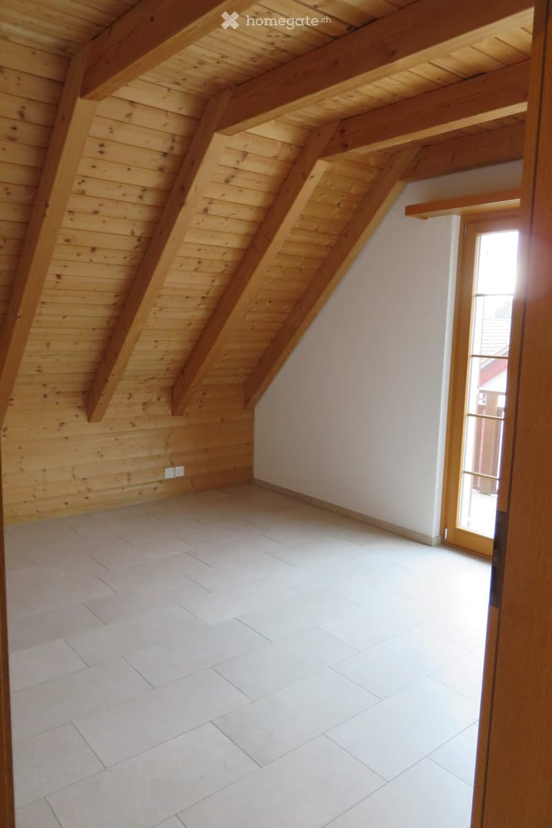 Empty attic space with wooden beams and slanted wooden ceiling, white tiled floor, and a window letting in natural light.