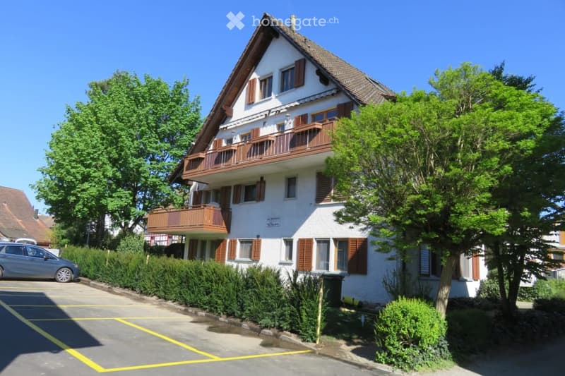 Two-story house with white walls, brown shutters, and balconies. The house has a well-maintained garden with trees and shrubs, and a parking area is visible in front.