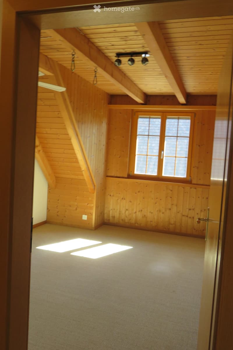Empty attic space with wooden walls, exposed wooden beams, and large windows allowing natural light to enter. The floor is carpeted, and the room has a high, slanted ceiling typical of attic spaces.