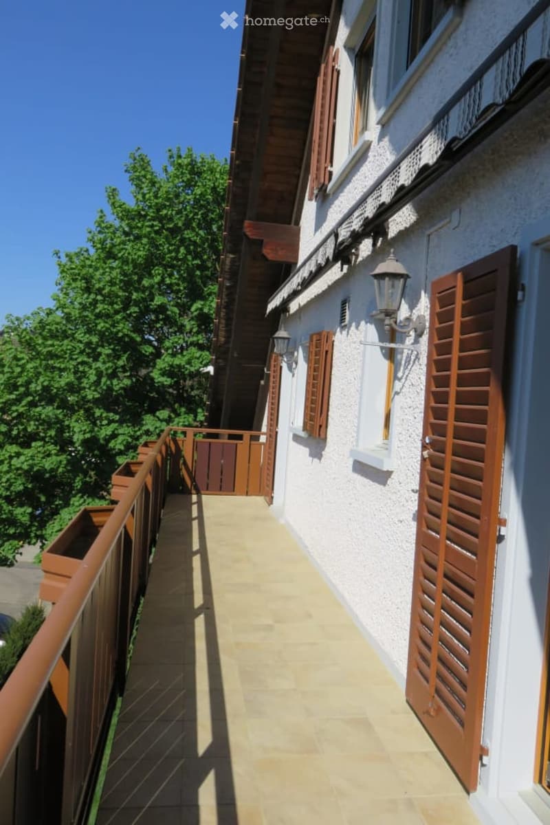 Balcony with wooden shutters, beige tiled floor, wooden railing, and small wooden gate leading to a garden area.