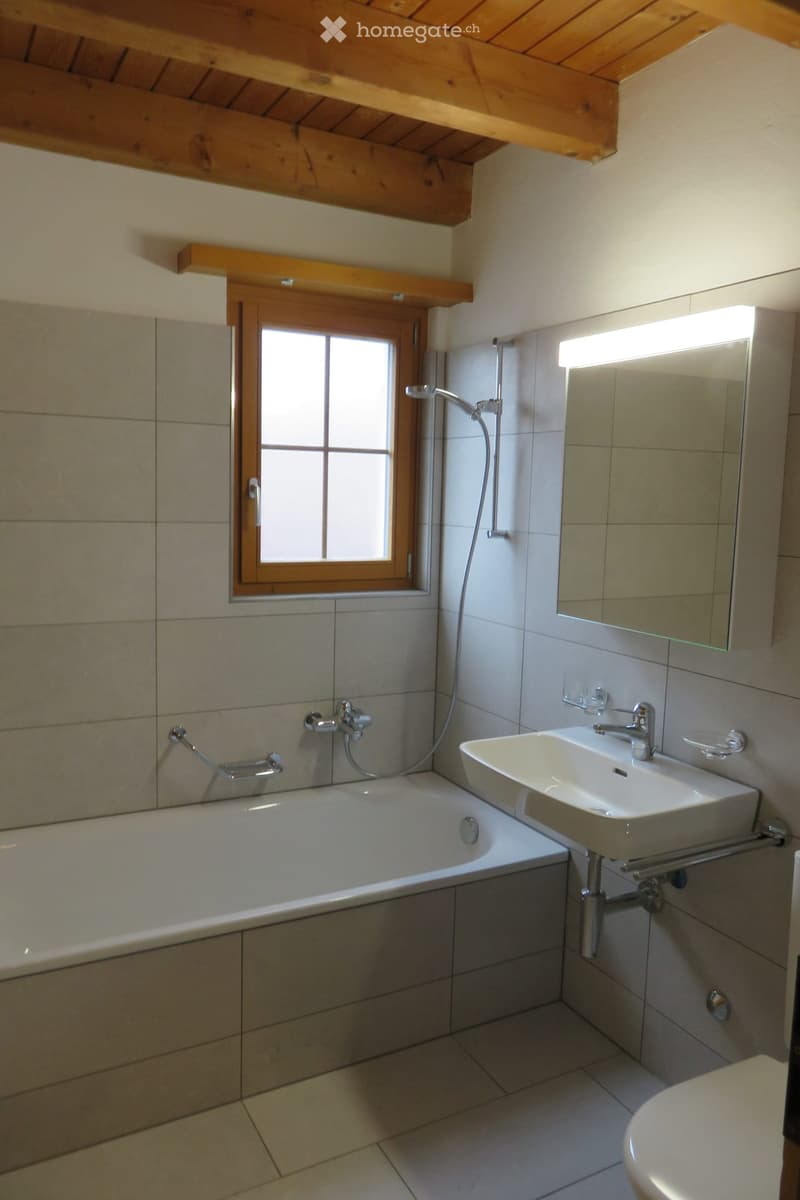 Modern bathroom with a bathtub, sink, mirror, and toilet. Features a wooden window frame and built-in wooden shelves above the window.