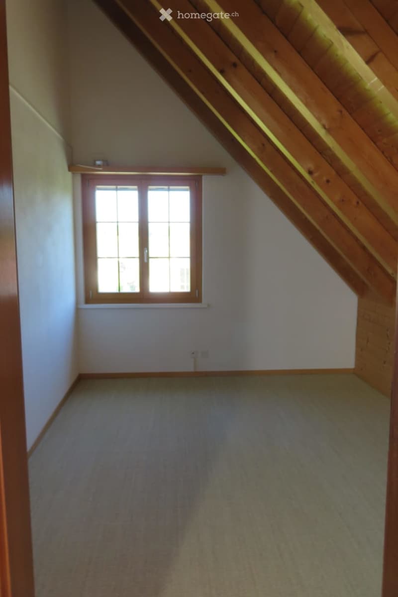 Empty attic room with wooden beams, carpeted floor, and a single window with wooden frame.