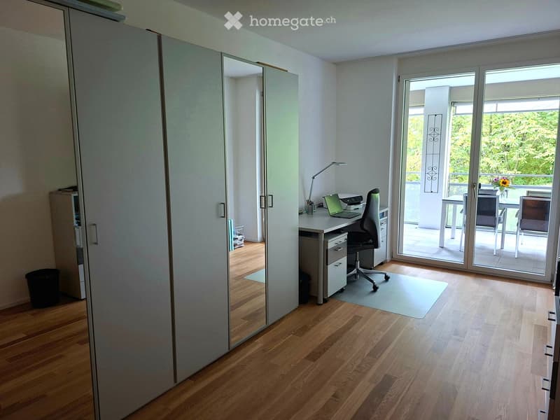 Bright, modern room with wooden flooring, a desk with a laptop and chair, a large mirror cabinet, and a sliding glass door leading to a balcony.