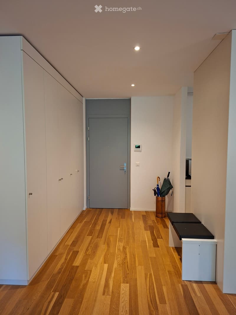 A narrow hallway with wooden flooring, white walls, and a grey door at the end. There is a small umbrella stand with umbrellas and a small bench on the right side.
