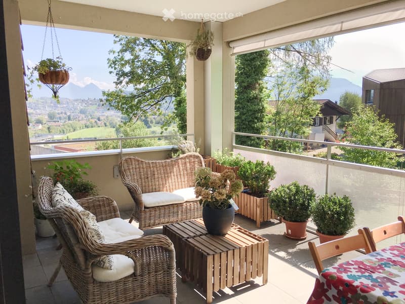 Balcony with wicker sofa, chairs, small table, and potted plants, overlooking a scenic valley with mountains.