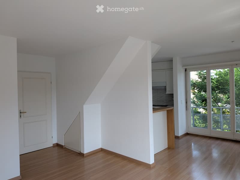 Empty room with wooden flooring, white walls, a door, and a small kitchen area visible in the background. Large windows and sliding doors lead to an outdoor area.