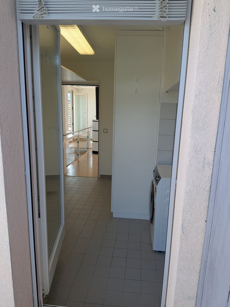 Hallway with tiled floor, white walls, and a washing machine in the corner. An open doorway leads to another room with wooden flooring and a staircase.