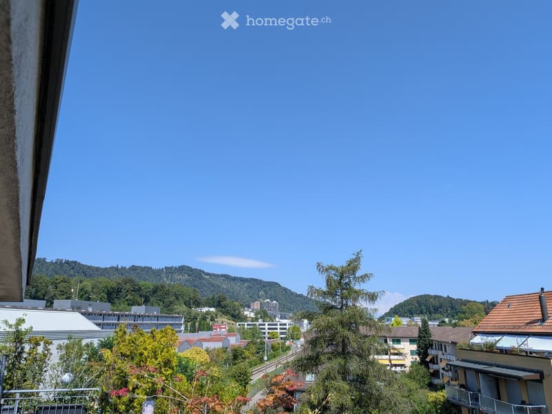 Balcony view overlooking a cityscape with buildings, trees, and distant hills under a clear blue sky.