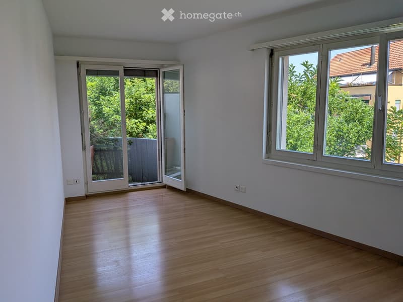 Empty room with wooden flooring, white walls, large windows, and a sliding glass door leading to a balcony.