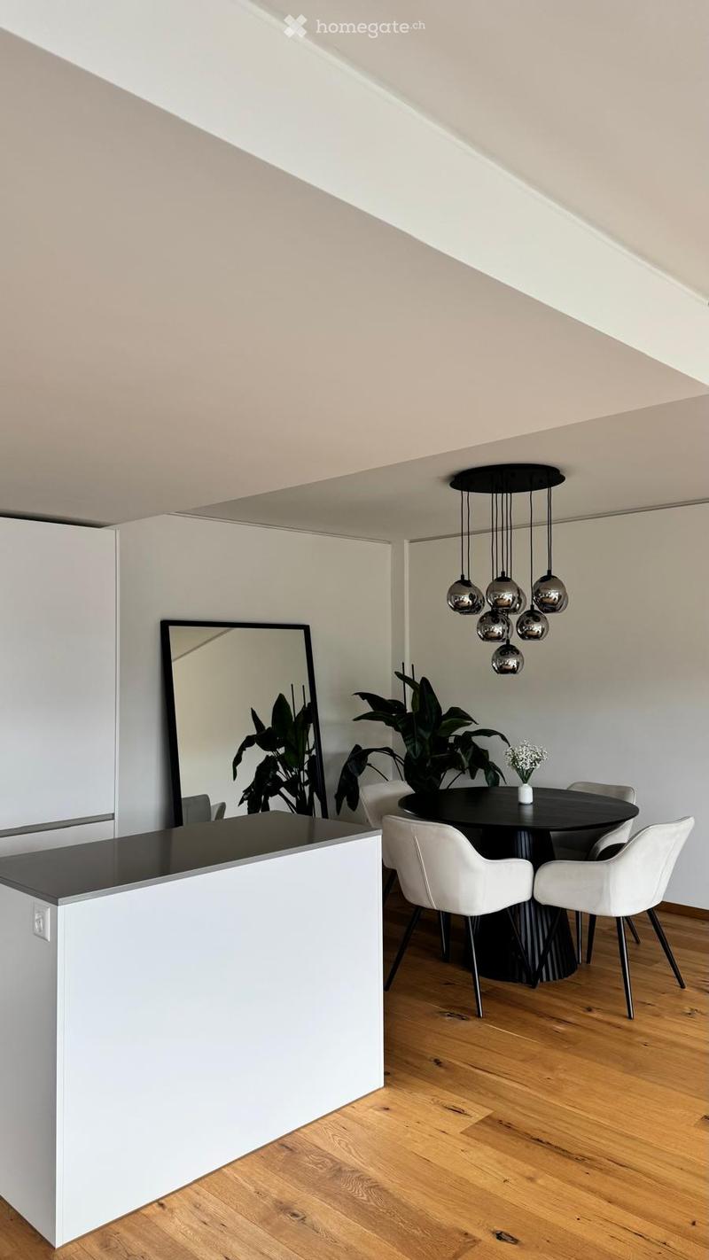 Modern dining area with a black round table, white chairs, a large mirror, and a hanging chandelier with spherical lights.