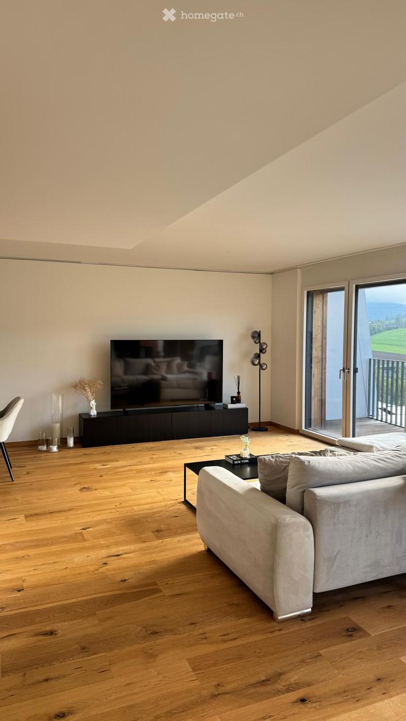 Modern living room with light wooden flooring, large windows offering a scenic view, a grey sofa, coffee table, TV, and decorative items.