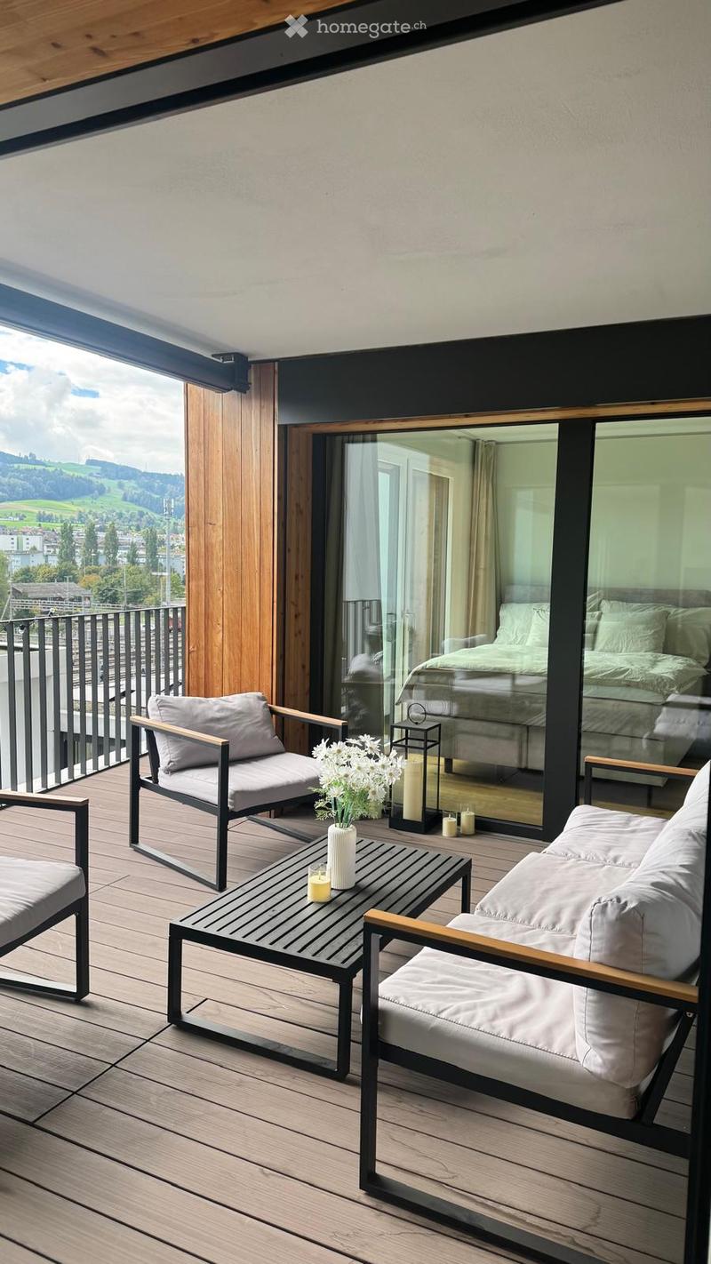Modern balcony with wooden decking, grey sofa, chairs, coffee table, vase with flowers, and candles, overlooking a scenic landscape.