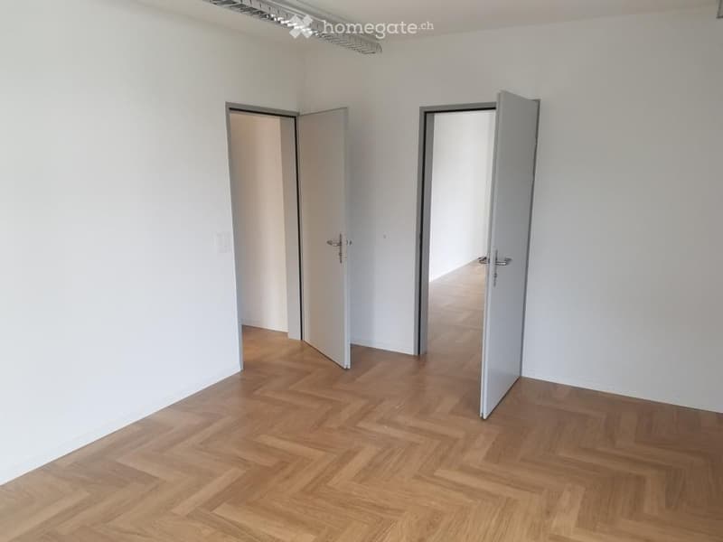 Empty room with light-colored walls and wooden flooring, featuring two open doors leading to adjacent spaces.