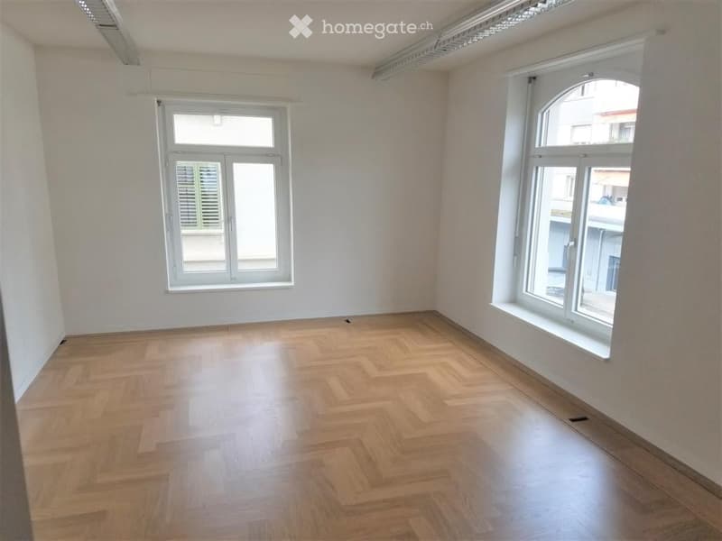 Empty room with wooden herringbone flooring, white walls, and large windows with shutters. No furniture or equipment present.