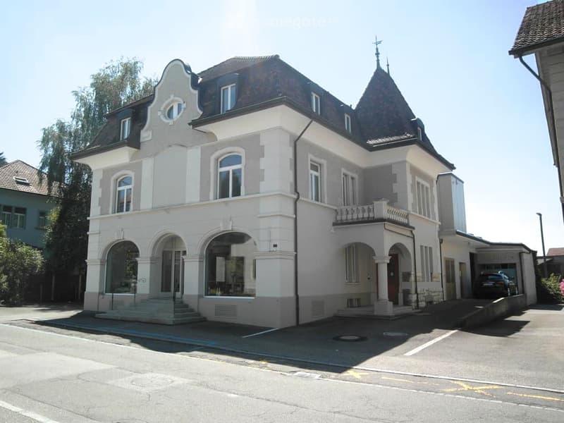 A large, multi-story building with a classic architectural style, featuring arched windows and doors, a prominent balcony, and a garage entrance.