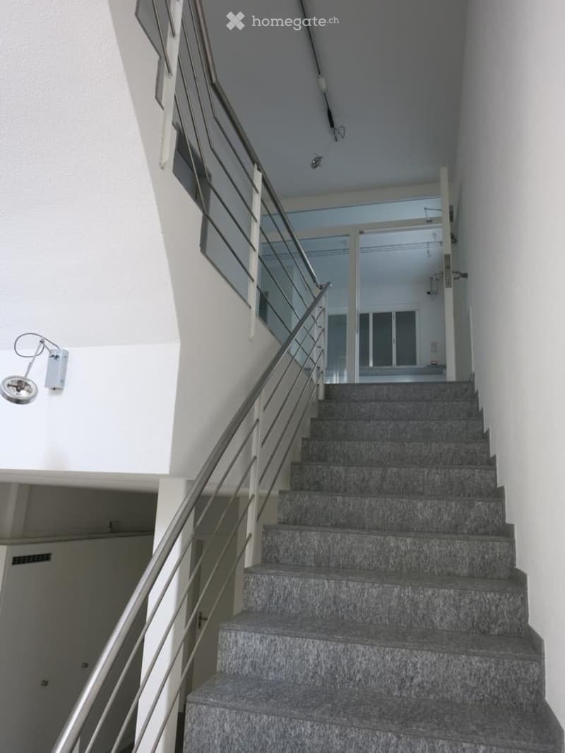 Modern interior staircase with grey marble steps and a metal railing, leading to an open area with large windows and bright lighting.