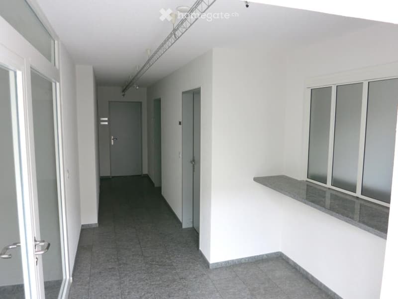 Empty hallway with tiled floor, white walls, and several doors. A small counter with windows is visible on the right side.