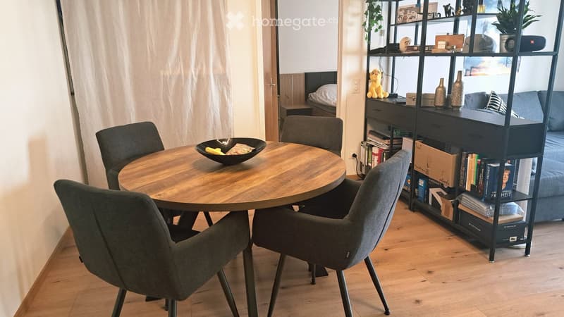 A modern, well-lit dining area with a round wooden table surrounded by four grey chairs. A tall black shelving unit holds books, vases, and decorative items. A small sofa with pillows is visible in the background, along with a plush toy on the shelf.