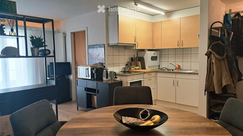A modern kitchen and dining area with light wooden cabinets, a microwave, oven, sink, and a dining table with chairs. There is a screen and a plant on a shelf, and a coat rack with jackets.