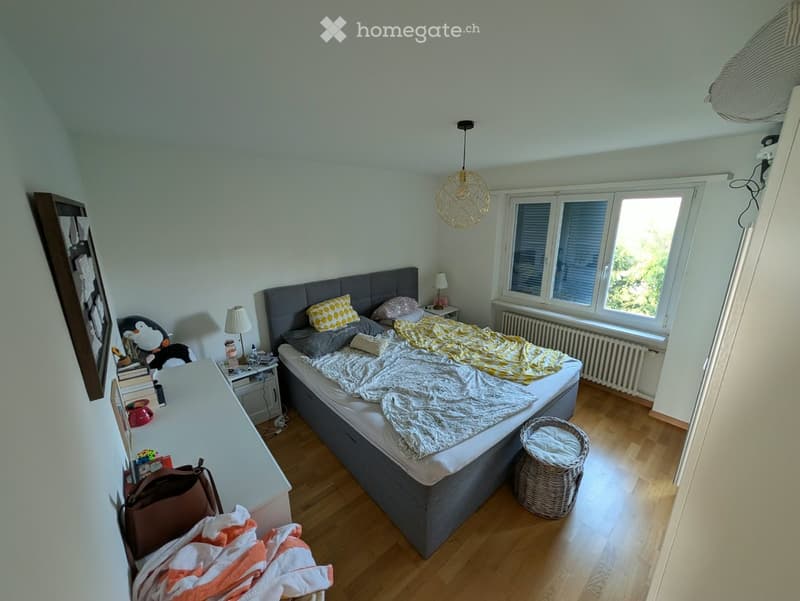 Cozy bedroom with wooden flooring, large window, grey upholstered bed, nightstand with lamp, wicker basket, and decorative items.