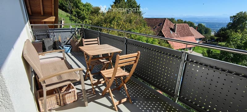 Balcony with a wooden table, two wooden chairs, and a wicker chair, overlooking a scenic landscape with trees and houses.