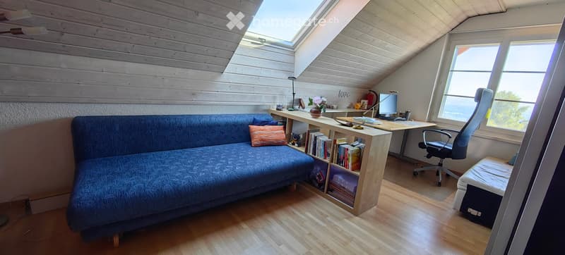 Cozy attic room with wooden ceiling, skylight, large window with ocean view, blue sofa, wooden desk, bookshelf, and office chair.