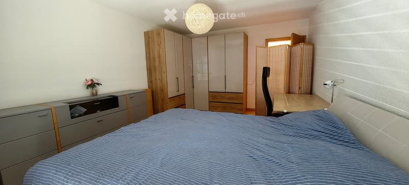 Modern bedroom with a blue bedspread, grey wardrobe, wooden desk, and a chair. Features a decorative flower vase on a side table and a hanging spherical light fixture.