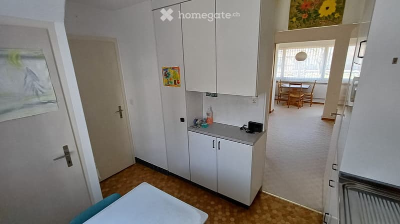 A small kitchen area with white cabinets, a countertop, and a sink, leading to a dining area with a table and chairs.