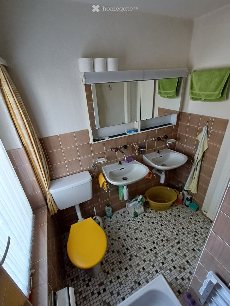 Bathroom with a toilet, sink, mirror, and bath. Brown tiled walls and a checkered floor. Toilet paper rolls on a shelf above the mirror. Towels hang on the wall. Cleaning supplies and a yellow bucket are on the floor.