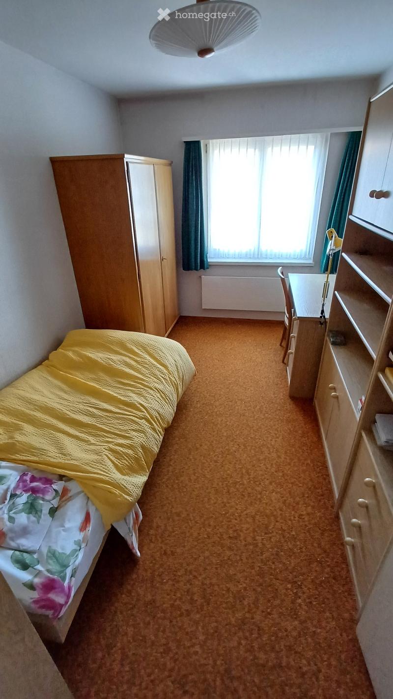 A small bedroom with a single bed, a wardrobe, a chair, a table, and a shelf. The room has carpeted flooring and a window with green curtains.