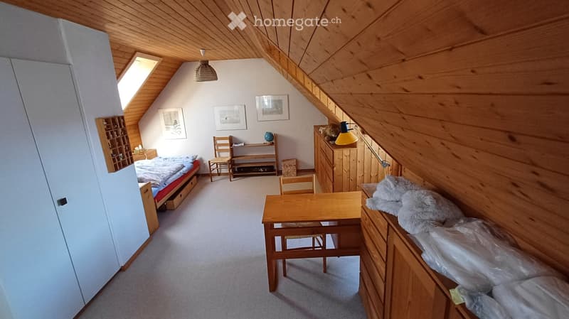 Cozy attic bedroom with wooden walls and ceiling, featuring a bed, a small table with chairs, a wardrobe, shelves, and artwork on the walls.