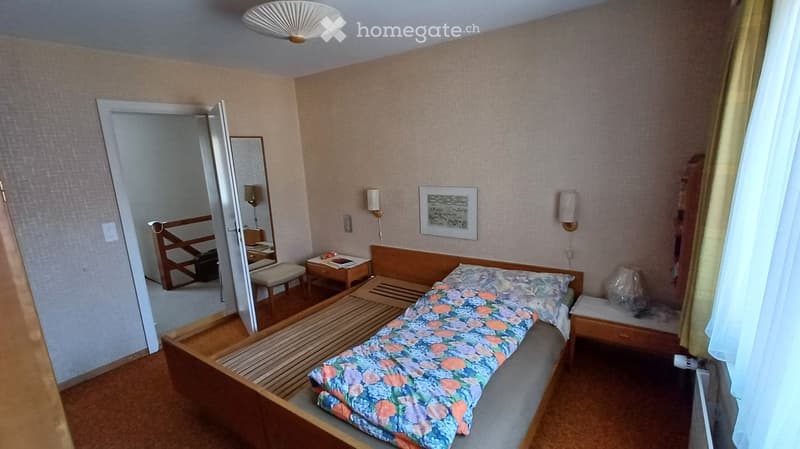 Cozy bedroom with a single bed, nightstand, mirror, and a small stool. The room features light-colored walls, wooden furniture, and a ceiling fan.