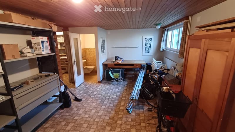 The room has a tiled floor, wooden ceiling, and includes a table with chairs, a bicycle, a stroller, shelves, and a visible bathroom area with a toilet and mirror.