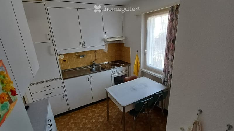 A small kitchen with white cabinets, a sink, stove, and oven. A small table with chairs is placed near the window.