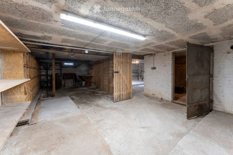 Empty basement space with concrete floors, exposed wooden beams, and unfinished walls. Features include a large open area, a partially enclosed section with wooden panels, and a small doorway leading to another area.