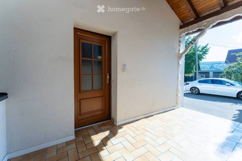Entrance area with a wooden door, tiled floor, and a covered walkway leading to a street with a parked car.