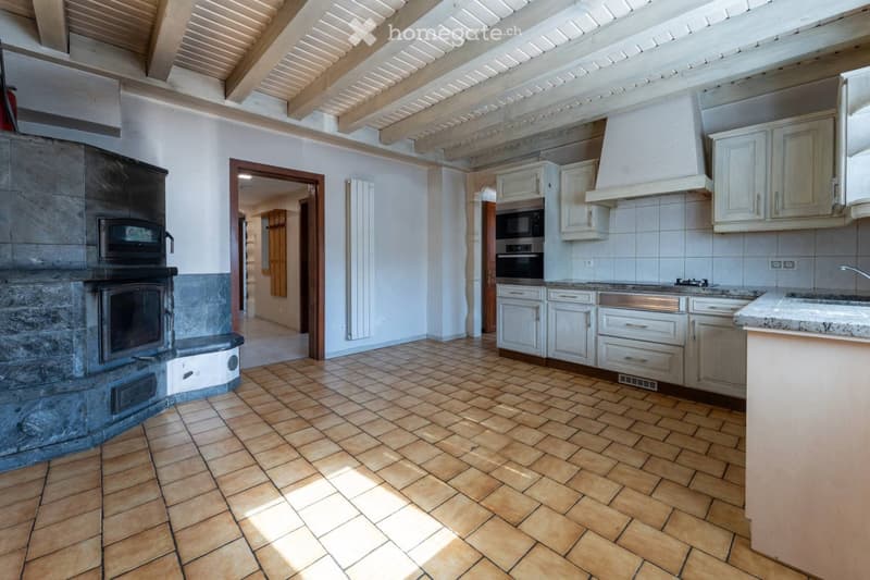 Open-plan space with a fireplace on the left, kitchen area on the right, and exposed wooden beams on the ceiling.