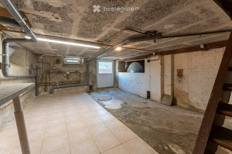 Empty basement with exposed pipes, concrete walls, tiled floor, and a wooden staircase leading upwards.